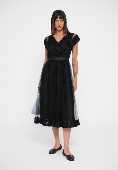 Rochie neagră cu dantelă florală, suprapune din tul, decolteu în V și mâneci scurte. Accentuata cu un brâu din satin și pantofi negri cu talpă plată.