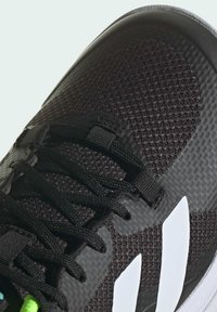 Svart sportskor med en strukturerad överdel i mesh-material, helt svarta detaljer och vita ränder på sidan. Gröna snören ingår.