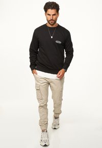 Black sweatshirt with logo at the chest, beige cargo pants with pockets, and sneakers featuring gray, white, and black accents.