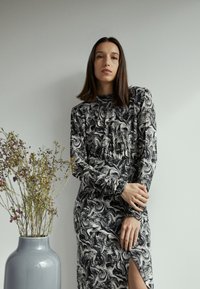 Vestido de manga longa em preto e branco com um decote alto, acompanhado por um vaso cinza contendo flores secas.