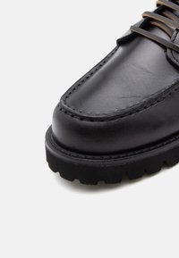 Chaussure en cuir noir avec un bout arrondi, des coutures visibles et une semelle en caoutchouc noir épais présentant un motif de traction. Des lacets bruns sont présents.