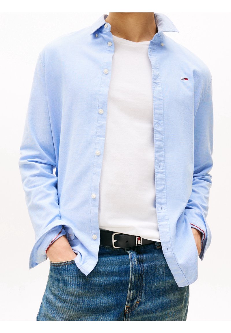 Homme portant une chemise bleu clair à boutons sur un t-shirt blanc, un jean bleu et une ceinture noire, les mains dans les poches, sur fond blanc.