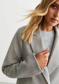 Ljust grått ullblandat ytterplagg med spetsig krage, med en subtil textur och överdimensionerade ärmar, knappstängning och rundad framkant.