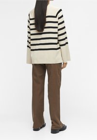 Gestreifter Pullover mit cremefarbener Basis und schwarzen horizontalen Streifen, kombiniert mit braunen Hosen. Verfügt über eine lockere Passform und lange Ärmel.
