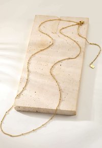 Goldene Kettenhalskette mit einem filigranen, strukturierten Design, auf einer hellen Steinoberfläche platziert. Sie verfügt über ein kleines Etikett in der Nähe des Verschlusses.