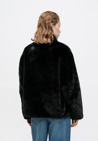 Chaqueta de pelo sintético negra con una textura suave, cuello alto y hombros caídos, combinada con jeans de color azul claro.