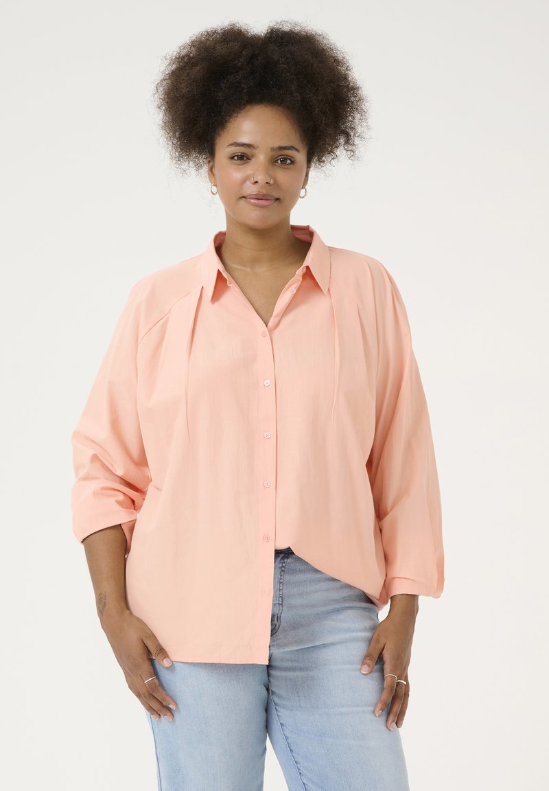 Camisa de botones de gran tamaño color durazno claro con cuello, diseño de hombros plisados y mangas remangadas, combinada con jeans de color azul claro.