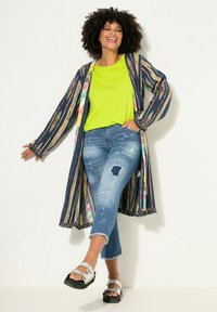 Kimono colorido y estampado con flecos, combinado con una blusa verde lima brillante y unos jeans claros desgastados, completado con sandalias plateadas.