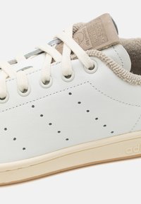 Biele kožené tenisky s šedými perforáciami, béžovými zamšanovými akcentmi, krémovými šnúrkami a štruktúrovanou gumovou podrážkou. Logo na jazyku.