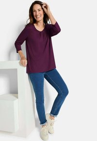 Lila Dreiviertelärmel-Oberteil mit einem subtilen Fältchen, kombiniert mit blauen Skinny-Jeans und weißen Sneakers, vor einem neutralen Hintergrund.