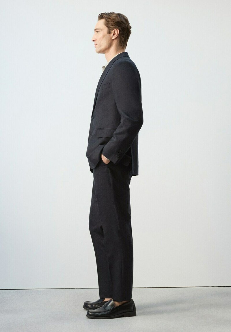 Homme debout de profil portant un costume sombre ajusté, les mains dans les poches et des mocassins noirs, sur un fond clair uni.