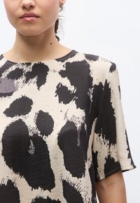 Επάνω μέρος σε ανοιχτό μπεζ χρώμα με αφηρημένο ζωικό μοτίβο σε μαύρο και γκρι, κοντά μανίκια και στρογγυλή λαιμόκοψη. Το υφασμάτινο υλικό με υφή προσθέτει βάθος.