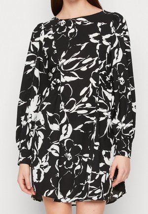 Femme portant une robe noire à manches longues avec des motifs floraux abstraits blancs et des épaules légèrement bouffantes.