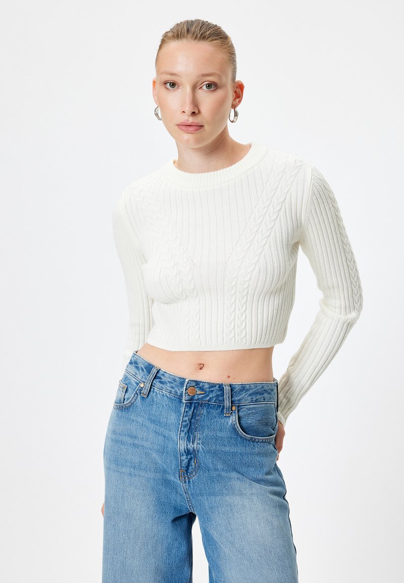 Maglione a coste bianco a maniche lunghe con design a treccia, abbinato a jeans azzurri a vita alta con tasche frontali.