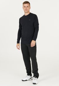 Långärmad svart skjorta med en subtil logo, kombinerad med svarta byxor och vita sportskor; enkel design, lätt tyg.