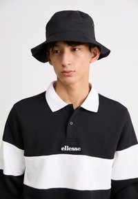 Juodai balta dryžuota ilgomis rankovėmis polo marškiniai su balta apykakle ir siuvinėtu logotipu. Modelis dėvi juodą kibirą.
