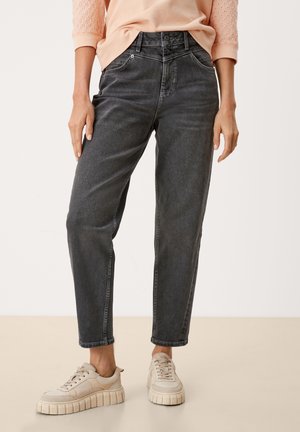 Vrouw draagt zwarte hoge taille jeans met taps toelopende pijpen, perzikroze top met kanten mouwen en beige plateau sneakers, staand op een neutrale vloer.