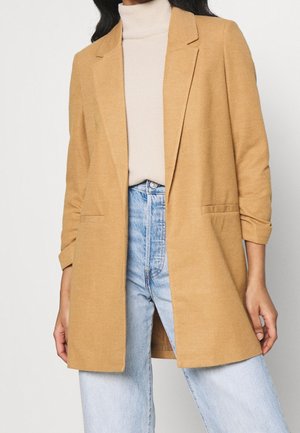 Vrouw draagt een camelkleurig open blazer over een crème coltrui en lichtblauwe high-waisted jeans.