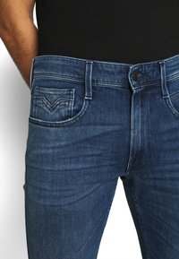 Vaqueros de mezclilla azul con corte slim, de altura media, con bolsillos delanteros y traseros, leve desgastado y cierre de botón en la cintura.