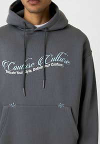 Grijze katoenen hoodie met trekkoorden en een voordezak. De lichtblauwe tekst luidt: "Couture Culture. Verhoog je stijl, bepaal je couture."