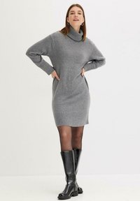 Robe pull en tricot grise avec un col roulé haut, des poignets et des ourlets côtelés, assortie à des bottes noires montantes. Texture lisse et silhouette ajustée.