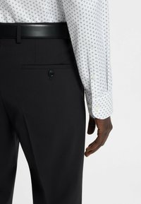 Pantaloni neri su misura con una texture elegante, abbinati a una camicia bianca con un motivo navy ripetuto. Cintura nera con una finitura liscia.