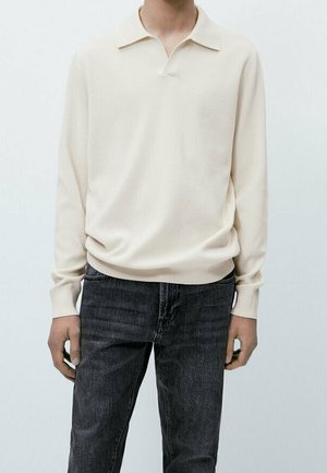 Man die een crèmekleurig poloshirt met lange mouwen en donkergrijze spijkerbroek draagt, staat voor een effen lichte achtergrond.