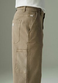 Pantalons cargo beiges à coupe droite, dotés de plusieurs poches et de détails de couture subtils. Fabriqués dans un tissu résistant avec une texture lisse.