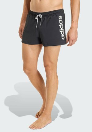 Man draagt zwarte Adidas zwemshort met witte trekkoord, staat op blote voeten tegen een lichte achtergrond.