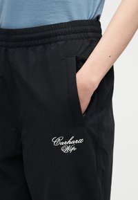 Άτομο που φορά μαύρο παντελόνι Carhartt Wip με το χέρι στην τσέπη και μπλε πουκάμισο ορατό στη μέση, πάνω σε απλό φόντο.