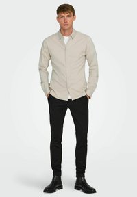 Camisa button-up de color beige claro con mangas largas, llevada sobre una camiseta blanca, combinada con pantalones negros ajustados y botas de tobillo negras.