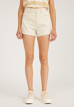 Femme portant un short en denim beige à ourlet roulé, un t-shirt rayé et des baskets beiges à plateforme, debout devant un fond clair uni.