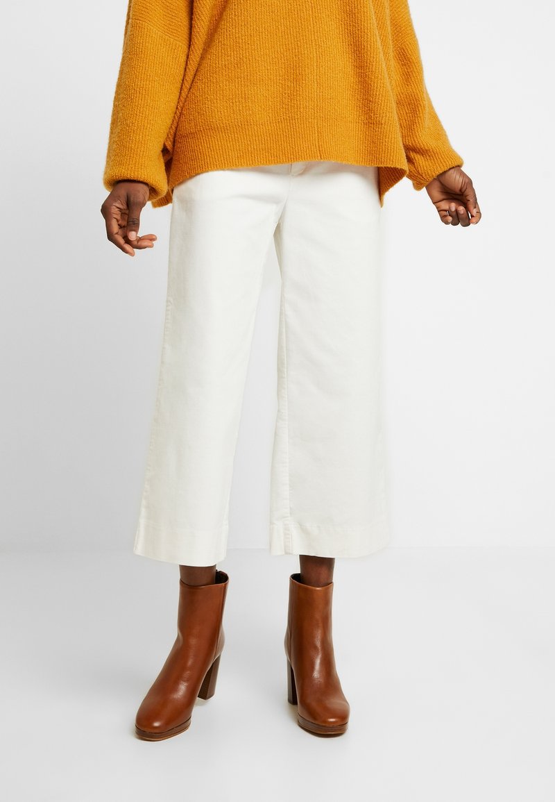 Överdimensionerad gul stickad tröja, krämfärgade vida byxor och bruna klackstövlar. Tröjan har en strukturerad stickning och byxorna har sidoflikar.