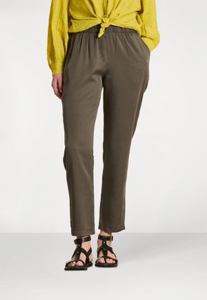 Personne portant un pantalon olive fuselé, des sandales noires à brides et une chemise jaune à manches longues nouée à la taille, debout sur un fond blanc.