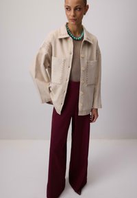 Beżowa kurtka zapinana na guziki z dwoma przednimi kieszeniami, haftowanymi detalami oraz szerokie czerwone spodnie, charakteryzujące się gładką teksturą i luźnym krojem.