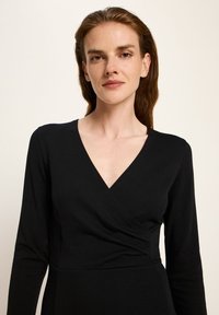 Frau mit langen braunen Haaren, die ein schwarzes Wickeloberteil mit langen Ärmeln vor einem schlichten hellen Hintergrund trägt.