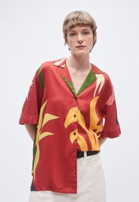 Camicia rossa a maniche corte con stampa botanica colorata in giallo e verde, con colletto a revers e chiusura frontale con bottoni.