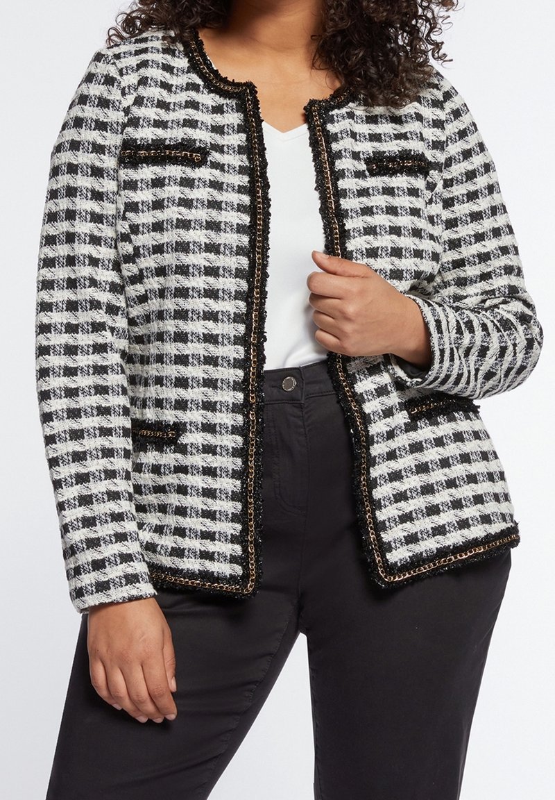Femme portant une veste texturée à carreaux noirs et blancs avec bordure dorée, haut blanc et pantalon noir, main tenant le revers de la veste.