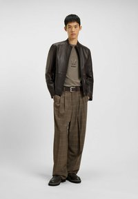 Donkerbruine leren jas over een groen T-shirt met een logo, gecombineerd met geruite bruine broek en zwarte schoenen. Eenvoudige riem en schone lijnen.