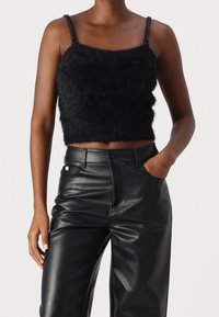 Top corto nero in pelliccia sintetica, con spalline sottili e finitura texture, abbinato a pantaloni in pelle nera a vita alta.
