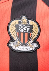 Maillot de sport rayé noir et rouge avec le blason du club OGC Nice montrant un aigle couronné et le texte "Depuis 1904."