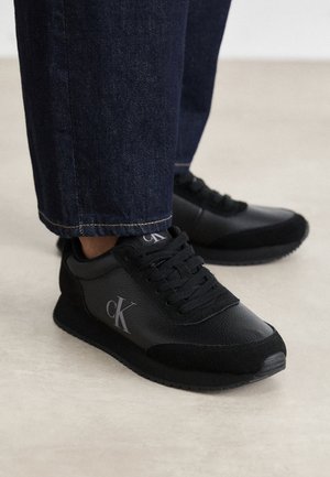 Czarne skórzane i zamszowe sneakersy z logo "cK" noszone z ciemnoniebieskimi dżinsami na jasnym tle podłogi.