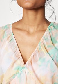 Blusa fluida en tonos pastel con escote en V, que presenta suaves patrones de tie-dye en verde, amarillo y rosa. Tejido ligero y aireado con detalles fruncidos.