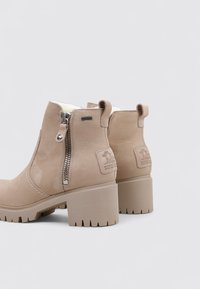 Botines beige con un acabado de cuero texturizado, cremalleras laterales y un tacón grueso. Interior forrado de felpa y suela de goma con tacos para mayor tracción.