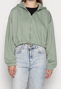 Sweat à capuche court vert menthe avec fermeture éclair à l'avant, cordon de serrage à la taille et poignets élastiqués. Associé à un jean taille haute bleu clair.