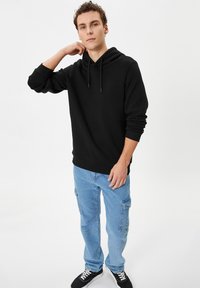 Zwarte capuchontrui met een kangoeroezak, gedragen met lichtblauwe cargobroek met meerdere zijzakken en zwarte sneakers.