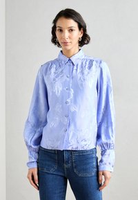 Lyselilla bluse med blomstermønster, knappet foran, spiss krage og lange puffermer. Brukt sammen med blå jeans. Glatt tekstur.