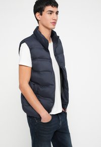 Granatowa pikowana kamizelka z wysokim kołnierzem, zamkiem błyskawicznym i dwoma bocznymi kieszeniami, noszona na białej koszulce, w zestawieniu z ciemnymi jeansami. Gładka faktura.