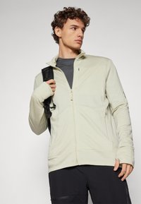 Light beige zip-up jacket made of soft fabric, featuring a high collar, thumbholes in the sleeves, and minimal branding.