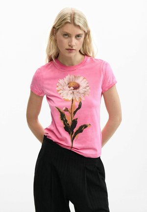 Jeune femme aux cheveux blonds portant un t-shirt rose avec un grand motif de marguerite blanche et un pantalon noir, posant les mains derrière le dos.